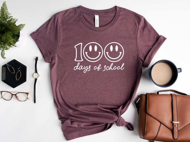 May include: Burgundy t-shirt with the text "100 days of school" and two smiley faces in white. The shirt is displayed on a white surface with accessories, including a watch, glasses, and a purse.