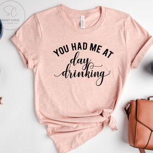 May include: A pink t-shirt with the text "You had me at day drinking" in black cursive lettering.