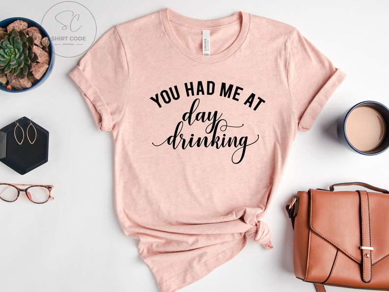 May include: A pink t-shirt with the text "You had me at day drinking" in black cursive lettering.