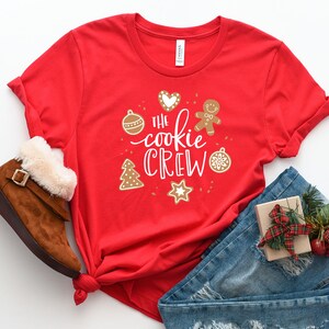 May include: Red t-shirt with a white graphic that says "The Cookie Crew" surrounded by Christmas-themed illustrations. The shirt is folded and laid on a white surface with a pair of brown boots, blue jeans, and a wrapped gift.