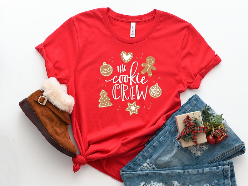 May include: Red t-shirt with a white graphic that says "The Cookie Crew" surrounded by Christmas-themed illustrations. The shirt is folded and laid on a white surface with a pair of brown boots, blue jeans, and a wrapped gift.