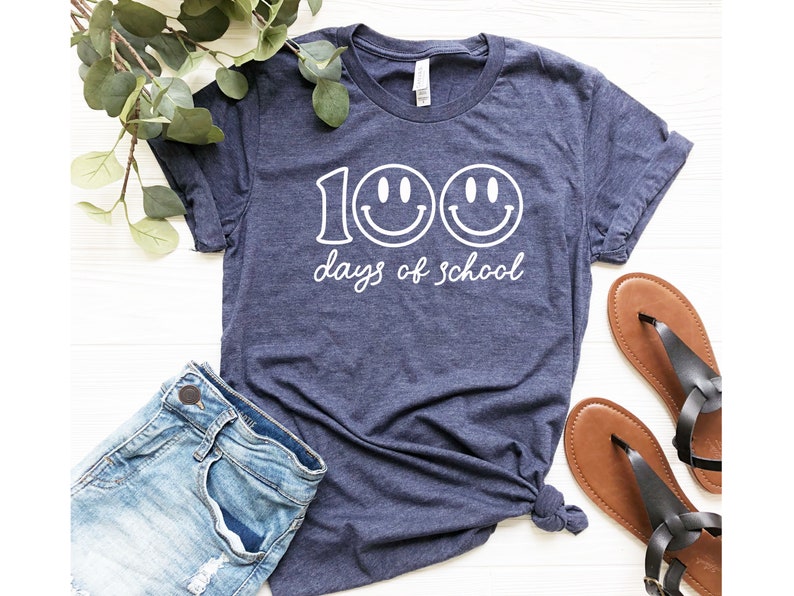 May include: Navy blue t-shirt with the text "100 days of school" and two smiley faces in white. The shirt is paired with blue denim shorts and brown sandals. Greenery is in the background.