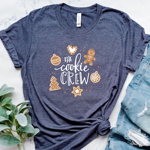 May include: A dark blue t-shirt with a white graphic that says "The cookie crew" surrounded by gingerbread cookies, a heart, and ornaments.