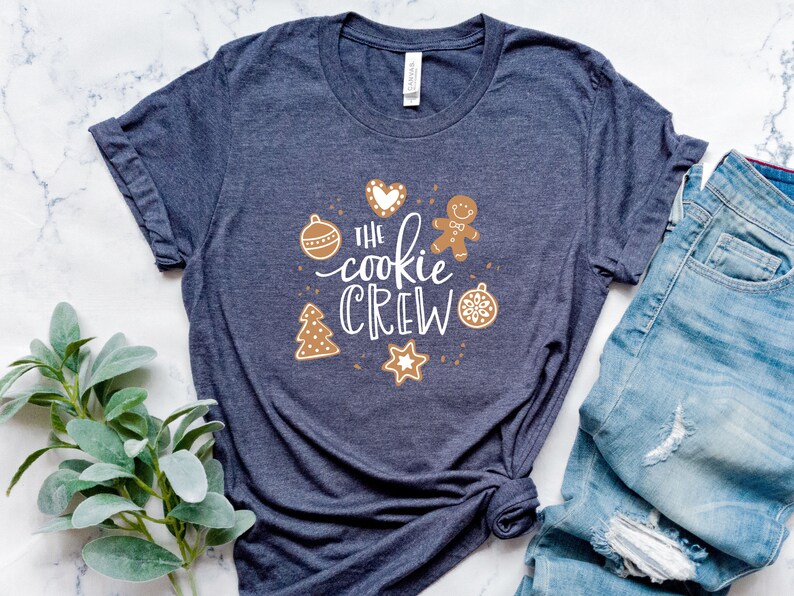 May include: A dark blue t-shirt with a white graphic that says "The cookie crew" surrounded by gingerbread cookies, a heart, and ornaments.