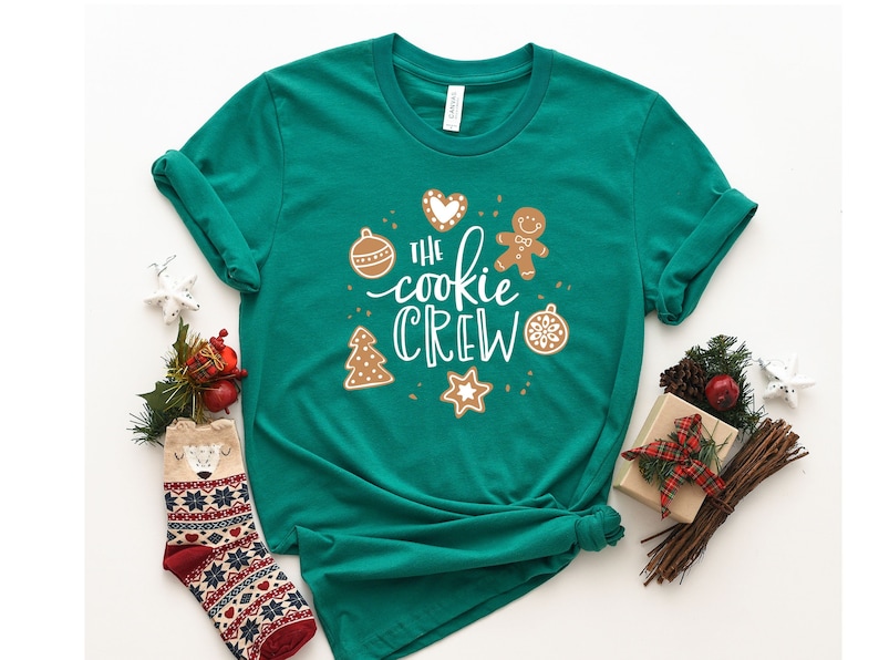 May include: A green t-shirt with the text "The Cookie Crew" in white lettering. The shirt features a gingerbread man, a Christmas tree, a star, and ornaments.