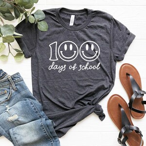 May include: Dark gray t-shirt with the text "100 days of school" and two smiley faces. The shirt is paired with blue jeans and brown sandals. The shirt is knotted at the waist.
