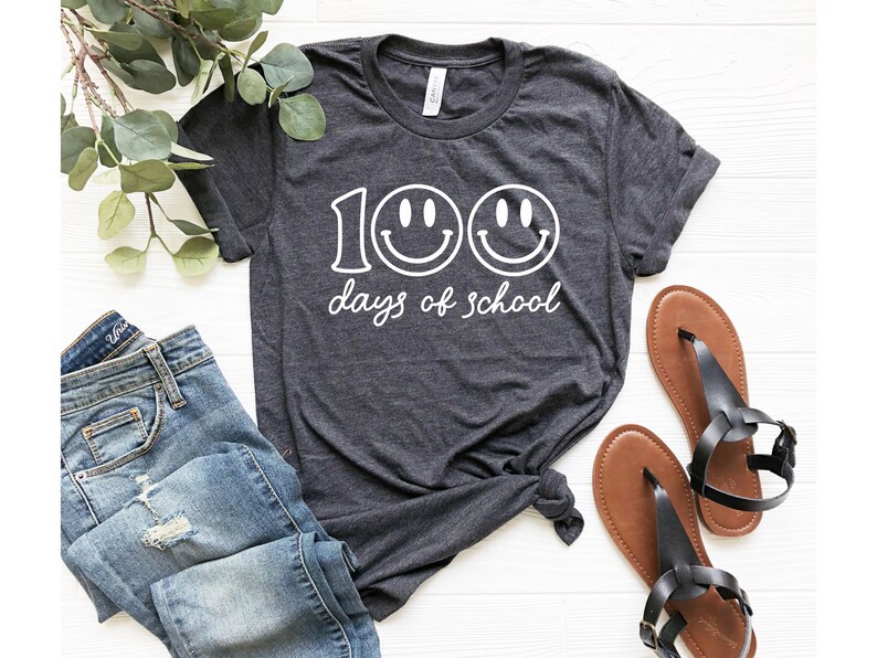 May include: Dark gray t-shirt with the text "100 days of school" and two smiley faces. The shirt is paired with blue jeans and brown sandals. The shirt is knotted at the waist.