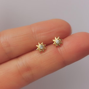 May include: A pair of gold-colored stud earrings, each featuring a sunburst design with a central, clear gemstone. The earrings are small and delicate, suitable for everyday wear. The earrings are displayed on a finger.