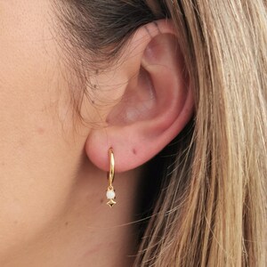 May include: Gold hoop earrings with a small white gemstone and a star-shaped charm. The earrings are dangling from a person's ear, with the focus on the jewelry. The earrings are a simple and elegant design.