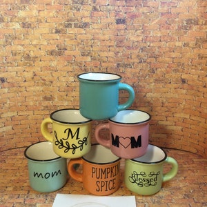 May include: A stack of six colorful ceramic mugs with black rims and handles. Each mug is a different color, including teal, yellow, pink, green, and orange. The mugs have various words and designs, such as "Mom", "M", "Pumpkin Spice", and "Blessed".