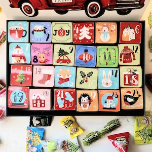 May include: Advent calendar with 24 numbered, illustrated squares in a black box. Each square features a different holiday-themed image, such as a snowman, Christmas tree, and a cozy sweater. Candy wrappers surround the box.