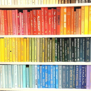 May include: A bookshelf filled with books arranged by color, creating a rainbow effect. The spines display titles and authors, with a variety of genres and sizes. The books are neatly organized on white shelves.