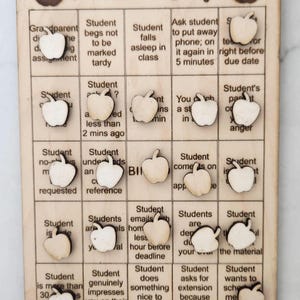 May include: Wooden Teacher's Bingo game with apple-shaped markers. The game board features various student behaviors and the text "Teacher's Bingo" at the top. A fun and engaging classroom activity.