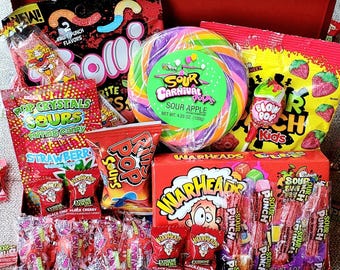 May include: A red box filled with a variety of sweets, such as a large swirl lollipop, bags of sour candies, and boxes of Warheads. The selection includes different flavours and brands, ideal for a sweet treat.