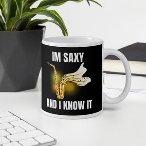 Puede incluir: Taza de cerámica blanca con fondo negro y el texto "IM SAXY AND I KNOW IT" en blanco. Un saxofón dorado con notas musicales está representado en la taza.