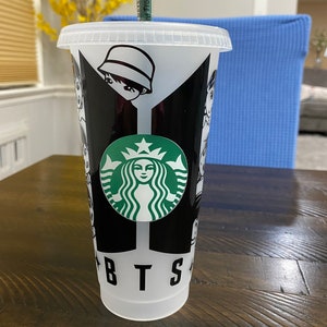 May include: A white reusable cup with a black and white design featuring the BTS logo and a Starbucks logo. The cup has a green straw.