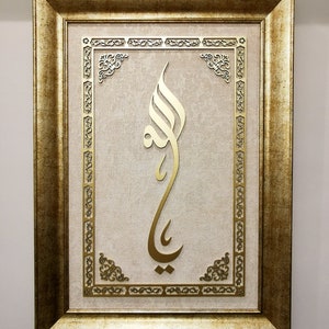 K&ouml;nnte beinhalten: Ein gerahmtes Kunstwerk mit einem goldenen arabischen Kalligraphie-Design auf beigefarbenem Hintergrund. Der Rahmen ist golden mit verzierten Details.