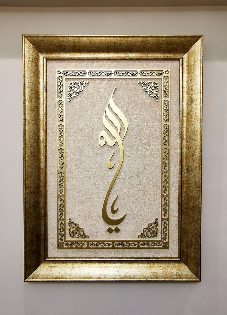 K&ouml;nnte beinhalten: Ein gerahmtes Kunstwerk mit einem goldenen arabischen Kalligraphie-Design auf beigefarbenem Hintergrund. Der Rahmen ist golden mit verzierten Details.
