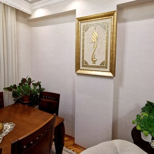 K&ouml;nnte beinhalten: Ein goldfarbener Bilderrahmen mit wei&szlig;em Hintergrund und goldener Kalligraphie. Das Bild h&auml;ngt an einer wei&szlig;en Wand in einem Raum mit einem Holztisch und St&uuml;hlen.
