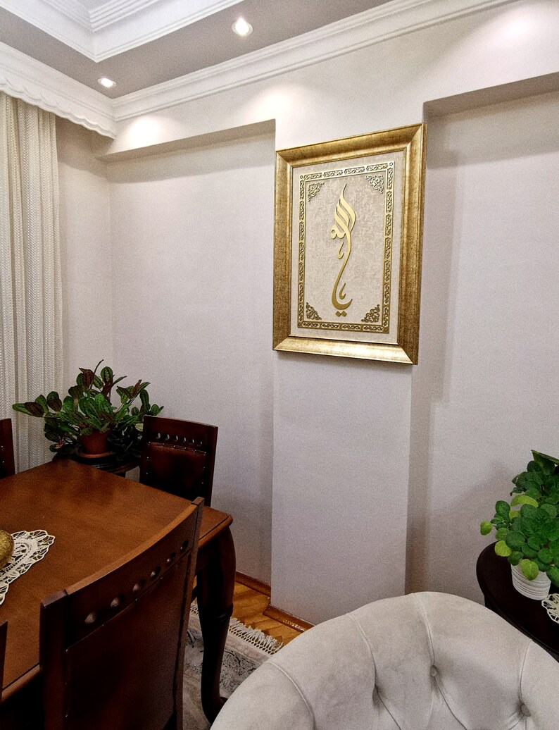 K&ouml;nnte beinhalten: Ein goldfarbener Bilderrahmen mit wei&szlig;em Hintergrund und goldener Kalligraphie. Das Bild h&auml;ngt an einer wei&szlig;en Wand in einem Raum mit einem Holztisch und St&uuml;hlen.
