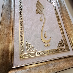 K&ouml;nnte beinhalten: Ein goldfarbenes gerahmtes Kunstwerk mit einem komplizierten Design. Das Kunstwerk zeigt eine dekorative, stilisierte arabische Kalligraphie in Gold auf einem hellrosa Hintergrund.