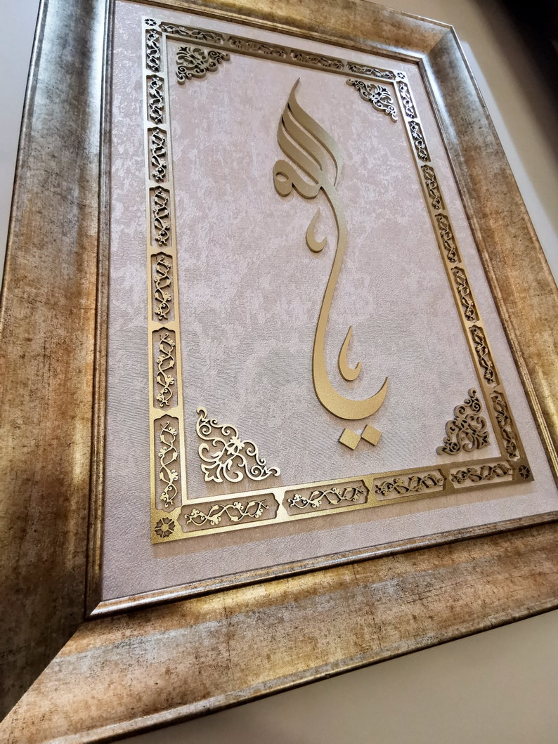 K&ouml;nnte beinhalten: Ein goldfarbenes gerahmtes Kunstwerk mit einem komplizierten Design. Das Kunstwerk zeigt eine dekorative, stilisierte arabische Kalligraphie in Gold auf einem hellrosa Hintergrund.