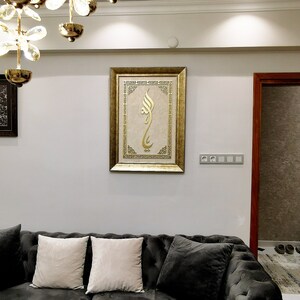 K&ouml;nnte beinhalten: Ein dunkelgraues Samtsofa mit zwei wei&szlig;en Kissen und einem goldgerahmten Bild mit islamischer Kalligraphie, das an der Wand dahinter h&auml;ngt.