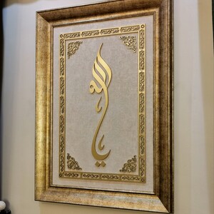 K&ouml;nnte beinhalten: Ein goldfarbener Bilderrahmen mit einem verzierten Design. Das Bild zeigt ein goldenes arabisches Kalligraphie-Design auf beigefarbenem Hintergrund.