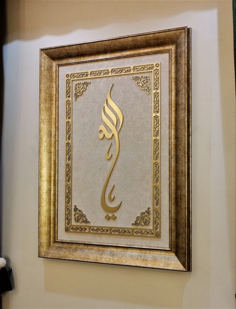 K&ouml;nnte beinhalten: Ein goldfarbener Bilderrahmen mit einem verzierten Design. Das Bild zeigt ein goldenes arabisches Kalligraphie-Design auf beigefarbenem Hintergrund.