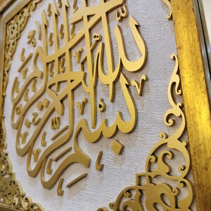 May include: A gold framed wall art piece with intricate carvings. The center of the art piece features a raised, gold Arabic calligraphy inscription.