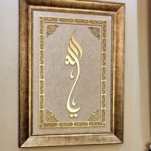 K&ouml;nnte beinhalten: Ein gerahmtes St&uuml;ck islamischer Kalligrafiekunst mit goldenen Buchstaben auf beigefarbenem Hintergrund. Der Rahmen ist verziert und hat eine goldene Oberfl&auml;che.