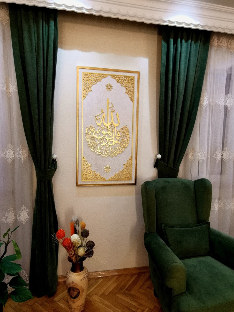 May include: A gold framed wall art piece with intricate designs and Arabic calligraphy. The art is hanging on a white wall with green curtains on either side.