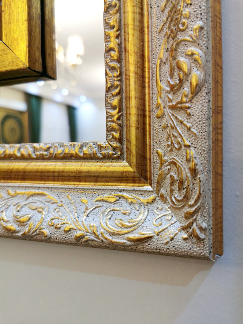 May include: Close-up of a gold and white ornate picture frame with a decorative floral design. The frame is made of wood and has a distressed finish.