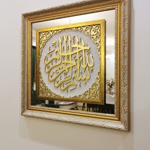 May include: A gold framed mirror with a white background and gold calligraphy inside. The calligraphy is in Arabic and reads "Allah".