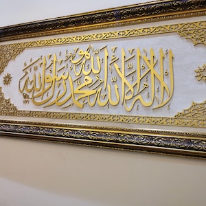 May include: A rectangular wall art piece featuring gold Arabic calligraphy on a white textured background. The artwork is enclosed in an ornate, dark brown and gold frame with intricate filigree patterns along the corners and borders.