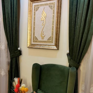 K&ouml;nnte beinhalten: Ein goldfarbener Bilderrahmen mit einem goldenen arabischen Kalligraphie-Design auf cremefarbenem Hintergrund. Das Bild h&auml;ngt an einer Wand mit gr&uuml;nen Vorh&auml;ngen auf beiden Seiten. Ein gr&uuml;ner Sessel mit einem floralen Kissen befindet sich im Vordergrund.