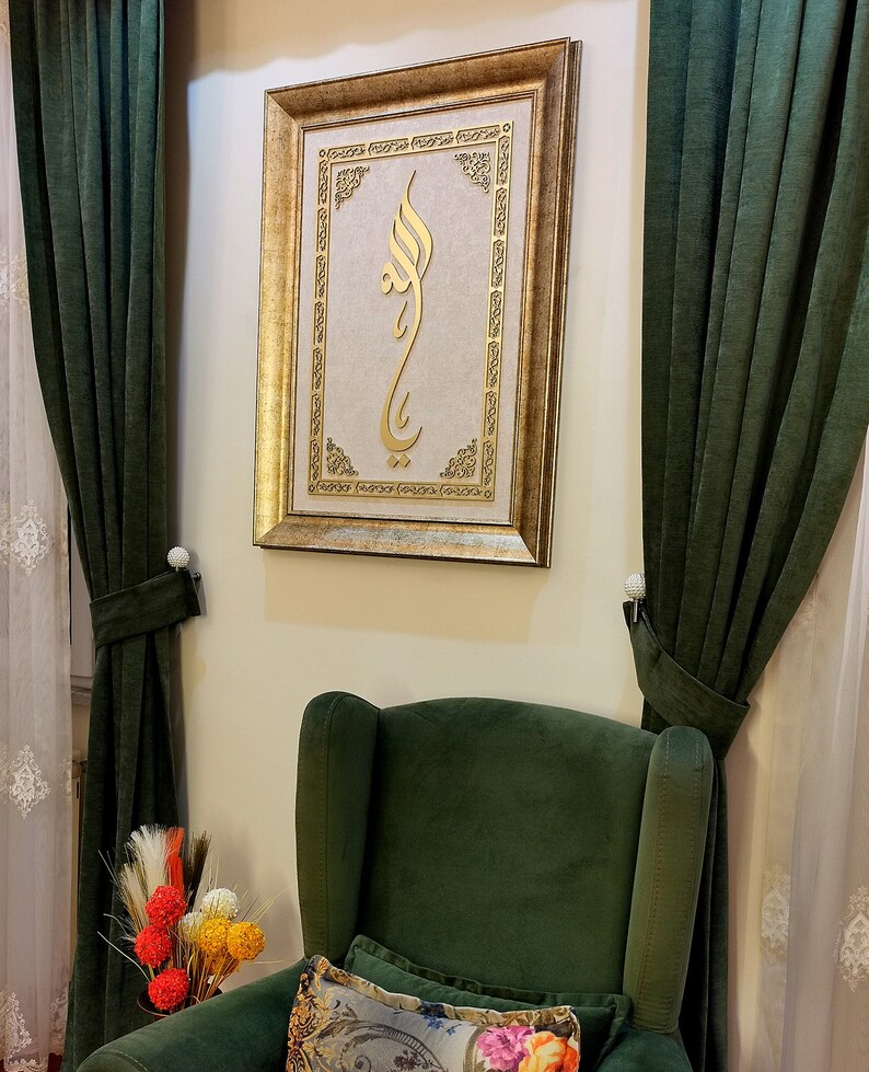 K&ouml;nnte beinhalten: Ein goldfarbener Bilderrahmen mit einem goldenen arabischen Kalligraphie-Design auf cremefarbenem Hintergrund. Das Bild h&auml;ngt an einer Wand mit gr&uuml;nen Vorh&auml;ngen auf beiden Seiten. Ein gr&uuml;ner Sessel mit einem floralen Kissen befindet sich im Vordergrund.