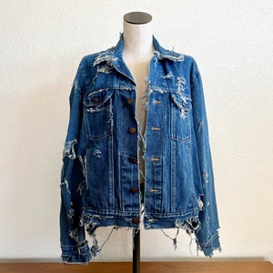 May include: A distressed blue denim jacket with a classic collar, button closure, and multiple pockets. The jacket features intentional rips and frayed edges, giving it a vintage, worn look. The jacket is displayed on a mannequin.