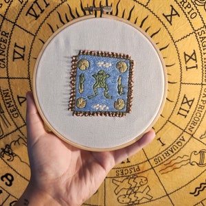 May include: A close-up of a hand holding a white embroidery hoop with a blue and green frog embroidered on a light blue fabric. The hoop is on top of a yellow and brown zodiac chart.