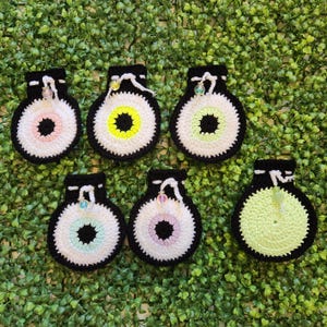 May include: Six crocheted eye-shaped pouches with different colored centers. The pouches are black with white trim and have a small bead at the top.