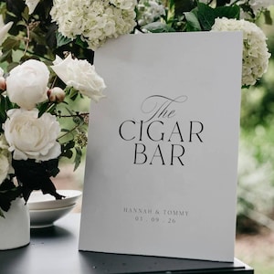 Può includere: Un cartello bianco con la scritta "The Cigar Bar" in elegante scrittura nera. Il cartello è appoggiato su una superficie nera, circondato da fiori bianchi e verde. Il cartello include anche i nomi "Hannah & Tommy" e la data "03.09.26".