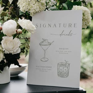 Peut inclure: Un panneau blanc avec l'inscription "SIGNATURE drinks" et des illustrations de cocktails. Le panneau est entouré de fleurs blanches dans un vase blanc. Les cocktails sont un Daiquiri et un Old Fashioned.