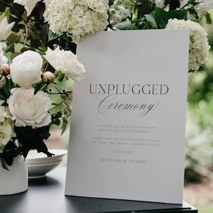 Puede incluir: Un letrero blanco con las palabras "UNPLUGGED Ceremony" en escritura negra sobre una superficie negra. Un arreglo floral blanco con hortensias y rosas está en un jarrón blanco. El texto invita a los invitados a estar presentes y apagar sus teléfonos.