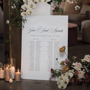 May include: A white seating plan with the words "Your Seat Awaits" and guest names, decorated with flowers and candles. The plan is for Veronica & Thomas's wedding on August 28, 2026. The setting is a venue with a rustic, elegant atmosphere.