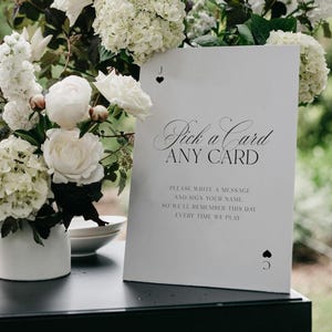 May include: A white rectangular sign with black text reading Pick a Card ANY CARD, followed by instructions to write a message and sign a name. The sign features a small black heart symbol at the top and a black spade symbol at the bottom.