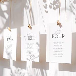Puede incluir: Tarjetas blancas con números de mesa con clips dorados colgadas de una viga de madera. Cada tarjeta está numerada y contiene una lista de nombres. Las tarjetas cuelgan sobre un fondo blanco, con adornos florales.