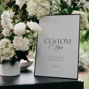 May include: A white sign with a black border displays the words "CUSTOM Sign" in elegant script. The sign is surrounded by white flowers and greenery, with a small white vase in the foreground. The sign also includes the text "FULLY EDITABLE" and initials.