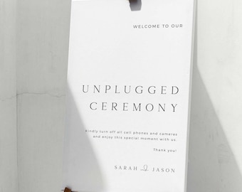 May include: White sign with the text "Welcome to our Unplugged Ceremony" and a request to turn off cell phones and cameras. The sign is on a wooden easel and the text is in a simple, modern font.