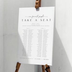 Peut inclure: Un plan de table blanc sur un chevalet en bois. Le texte "our favorite people TAKE A SEAT" est écrit en noir. Des noms sont listés sous des numéros de table dans une police claire. Les noms Claire et Henry sont en bas. Le chevalet est en bois.