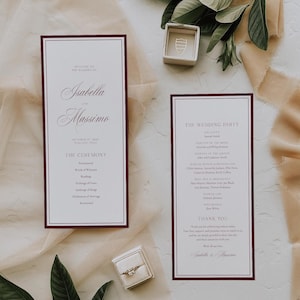 May include: Two rectangular wedding programs with white paper and dark burgundy borders. The text includes Isabella and Massimo, The Ceremony, and The Wedding Party. A small square ring box with a gold ring and a separate empty ring box are present.